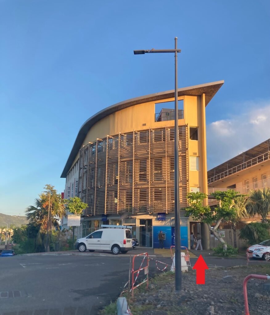 Vue extérieure d'un bâtiment moderne à la façade ocre et courbée, équipée de brise-soleil en bois. Une grande flèche rouge au premier plan indique le chemin vers l'entrée située sur le côté droit de l'immeuble.